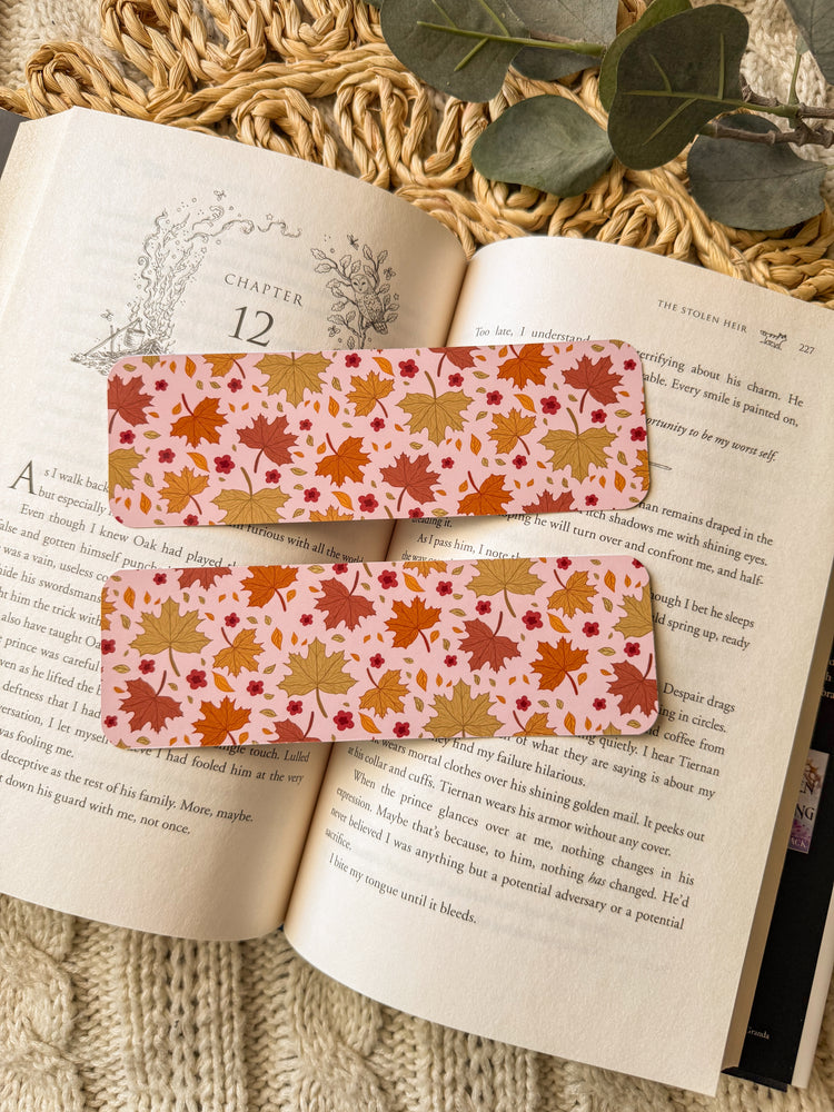 Two autumn-themed bookmarks on an open book with a textured surface and green leaves in the background.