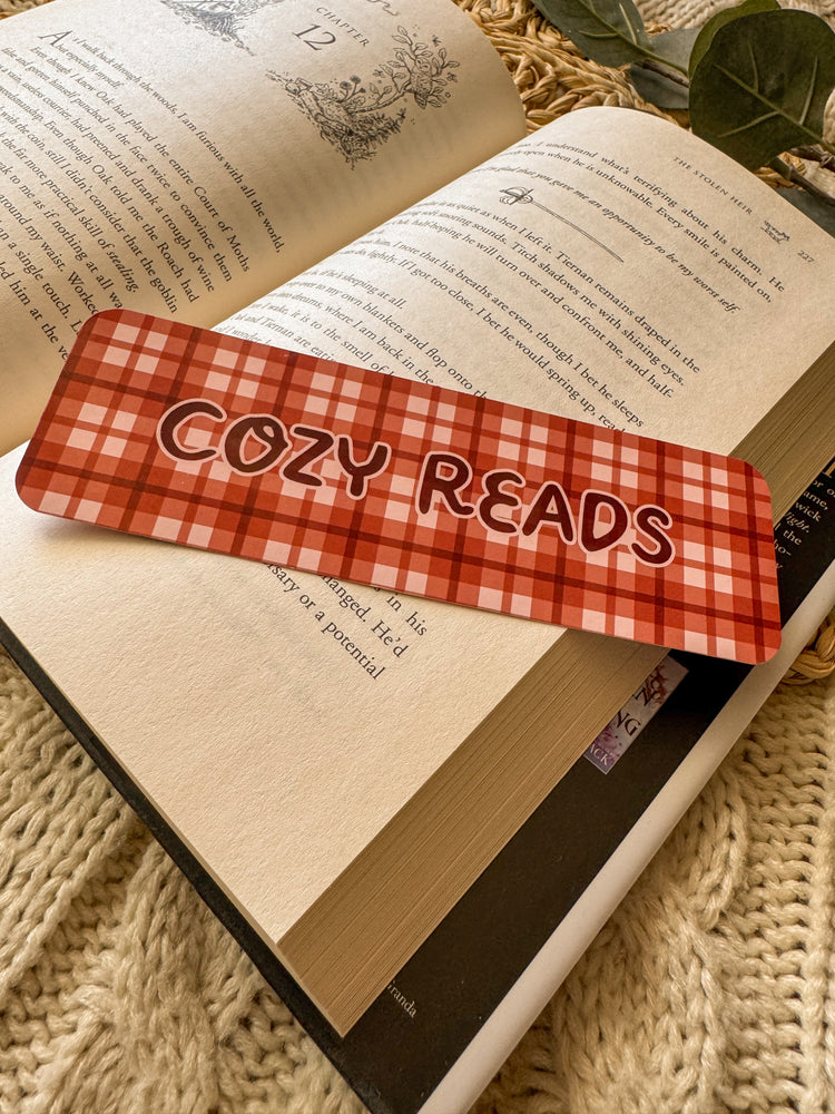 Open book with a maroon and white checkered bookmark labeled 'Cozy Reads' on a textured surface.