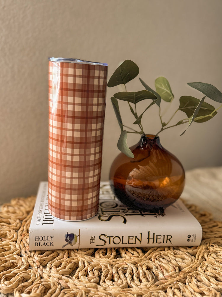 Plaid tumbler on a book with an amber glass vase and green leaves on a textured surface.
