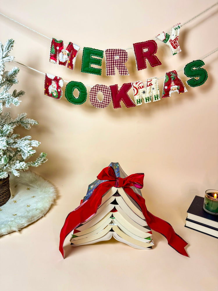 Handmade Merry Bookmas fabric banner hanging above a cozy holiday book scene with a book tree and candle.