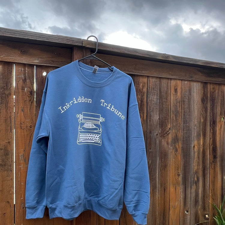 A blue sweatshirt with the text 'Inkridden Tribune' and an image of a typewriter on it, hanging on a wooden fence.