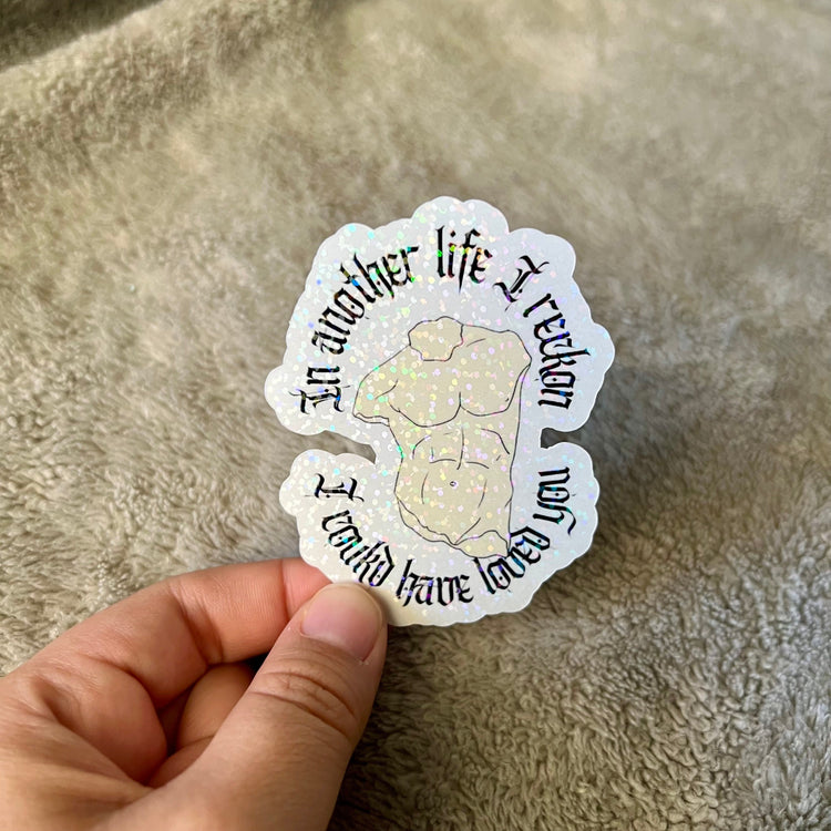 Sticker placed on a textured blanket, held between two fingers for cozy detail.