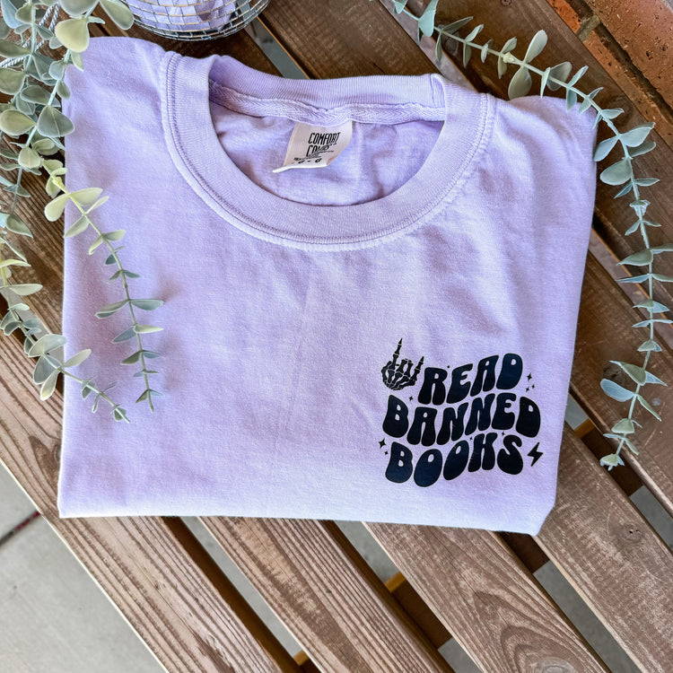 A lavender-colored T-shirt with the text 'Read Banned Books' printed on the front pocket, displayed on a wooden surface surrounded by foliage.