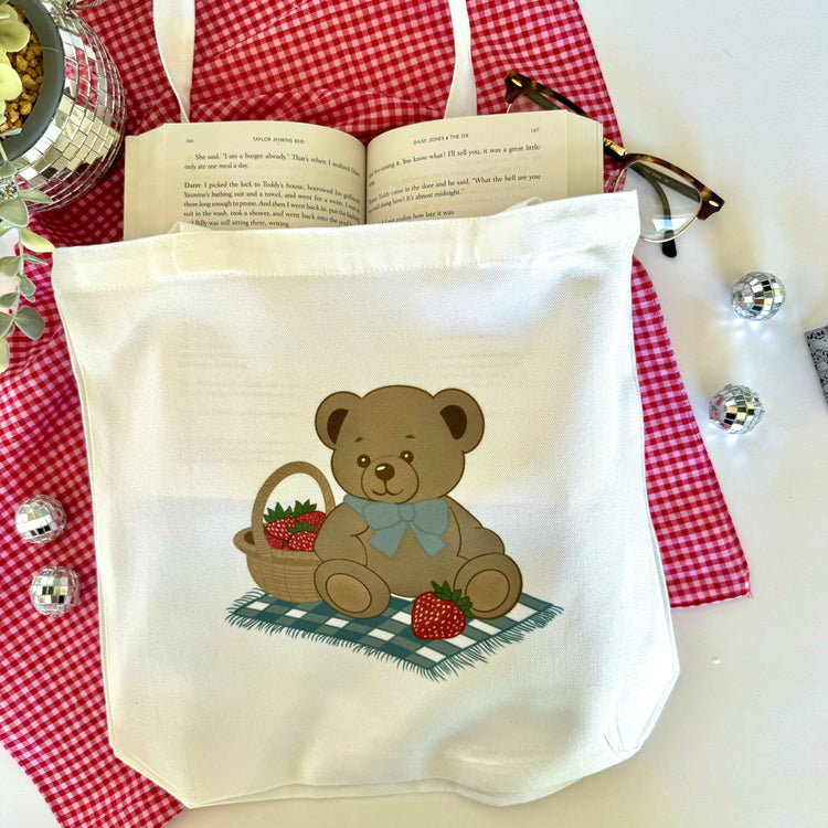 A white canvas tote bag with a teddy bear holding a basket of strawberries, displayed on a red and white checkered surface with a book and decorative items in the background.