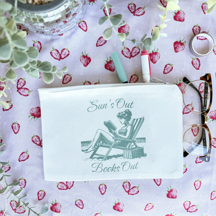 A white canvas pouch with a printed text 'Sun's Out Books Out' and an illustration of a person sitting on a chair. The pouch is placed on a surface with a floral pattern, and there are glasses and a pen beside it.