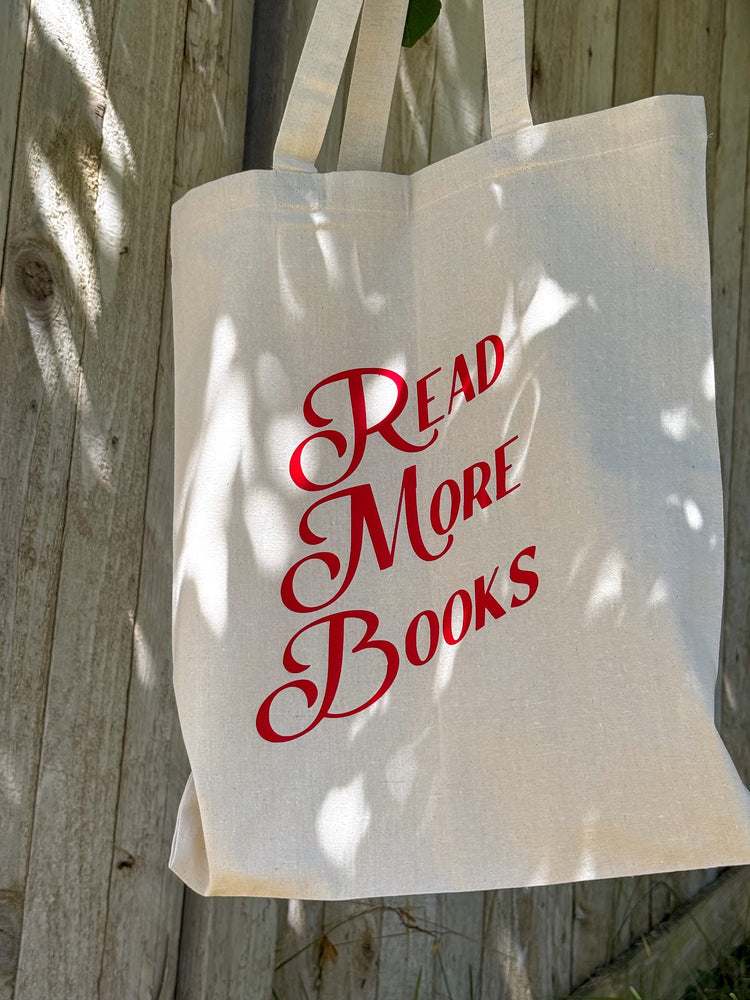 A beige canvas tote bag with 'READ MORE BOOKS' printed in red typography on the side, displayed against a wooden background.