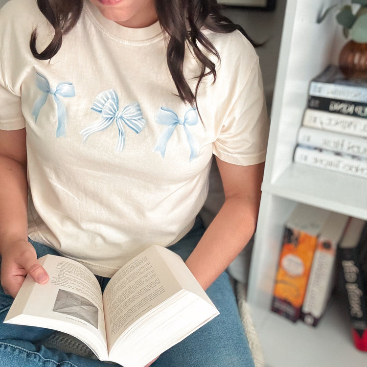 A person wearing a beige cropped t-shirt with blue bow graphics, sitting in front of a bookshelf and reading a book.