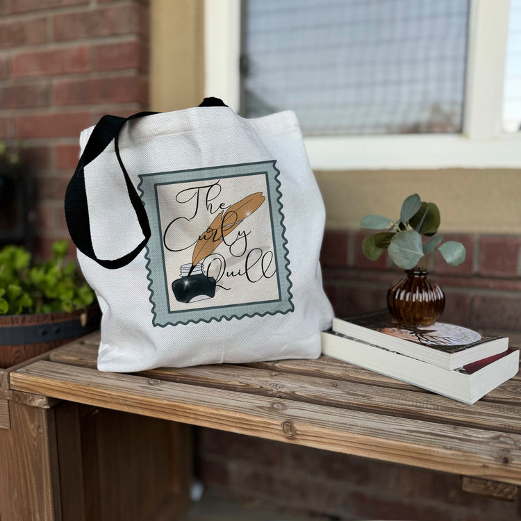 A white cotton tote bag with black straps featuring a printed design that includes a stylized representation of a book and a quote, placed on a wooden surface with a small plant and a book in the background.