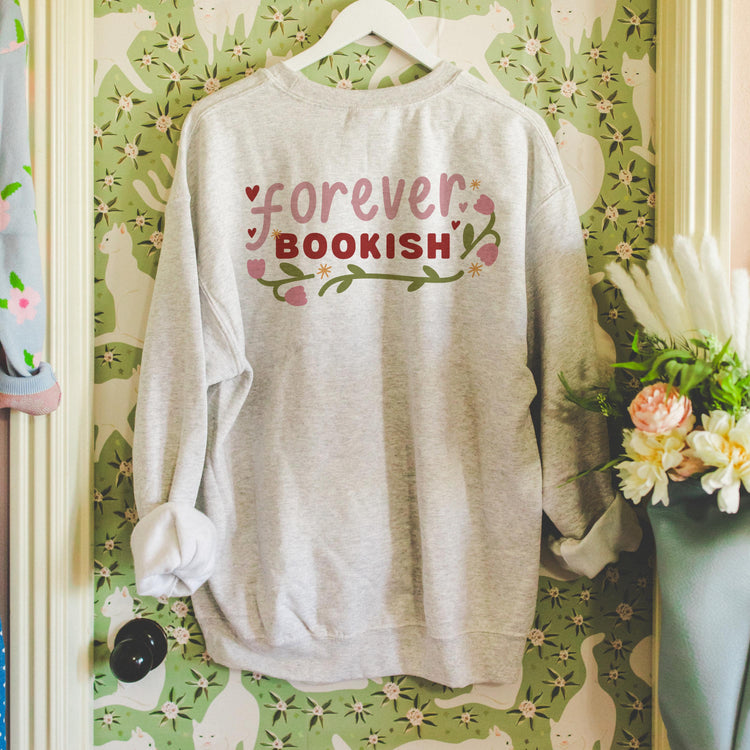 A gray sweatshirt with the text 'forever BOOKISH' printed on the front, featuring a floral design, displayed on a hanger against a wall with floral wallpaper.