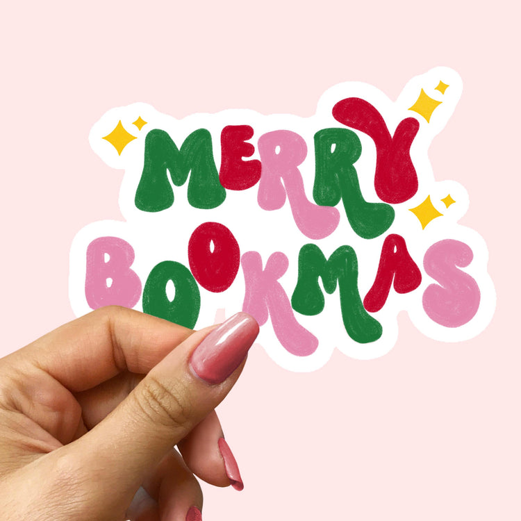 Hand holding a colorful 'Merry Bookmas' sticker against a pink background