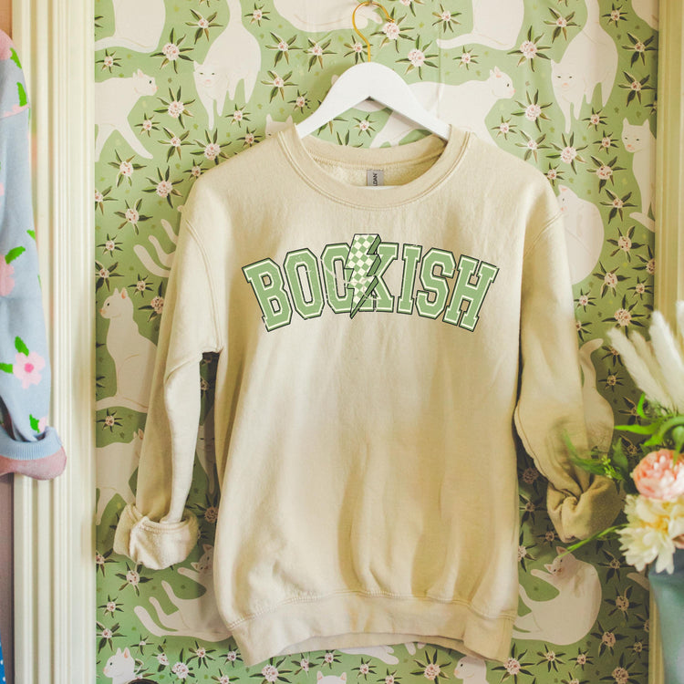 Beige Bookish Bolt sweatshirt with whimsical lightning/book graphic, styled with books and cozy reading props.