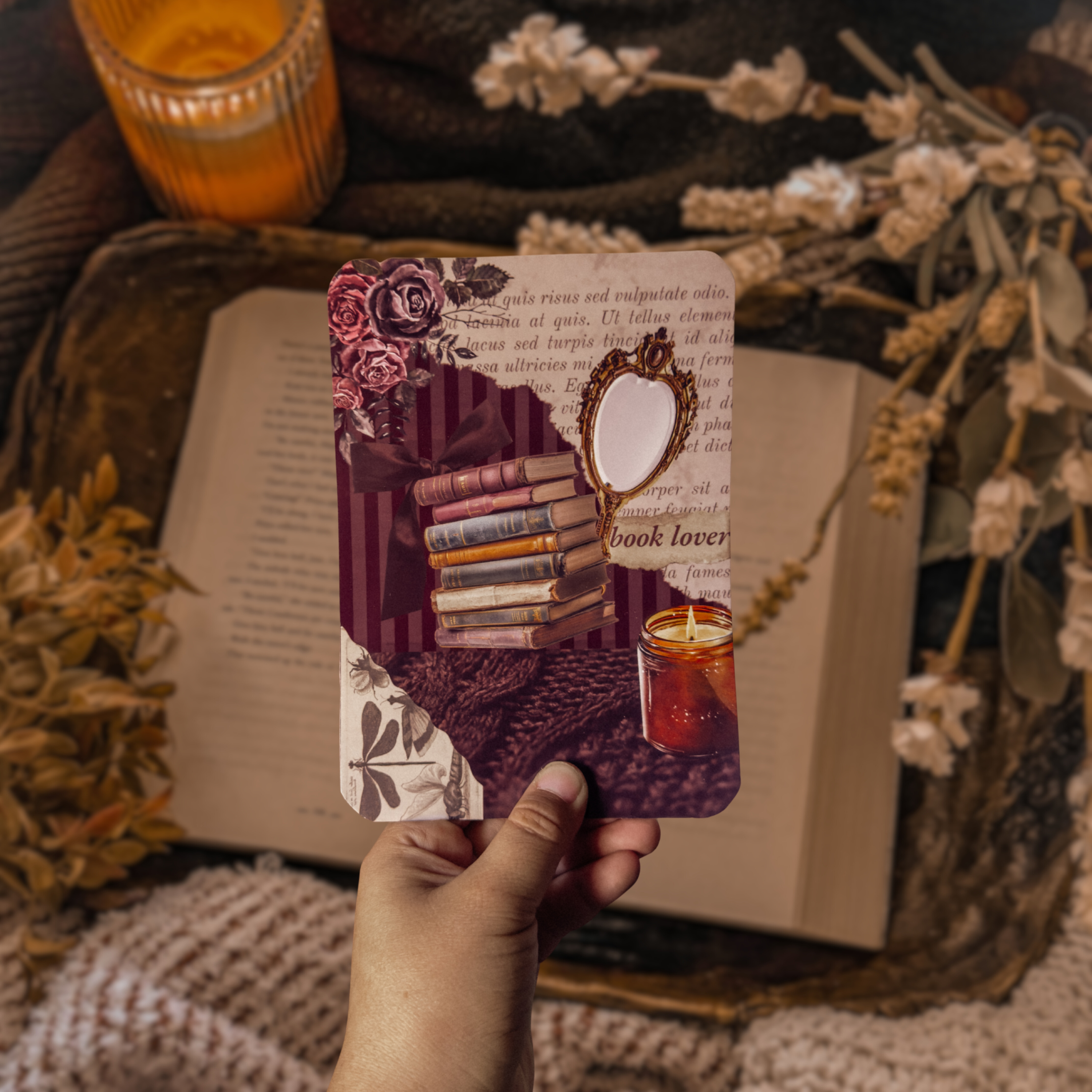 Lifestyle shot of a hand holding the Midnight Library Kindle insert, featuring a purple floral and vintage book collage, styled against an open book and dried flowers in a cozy library setting.