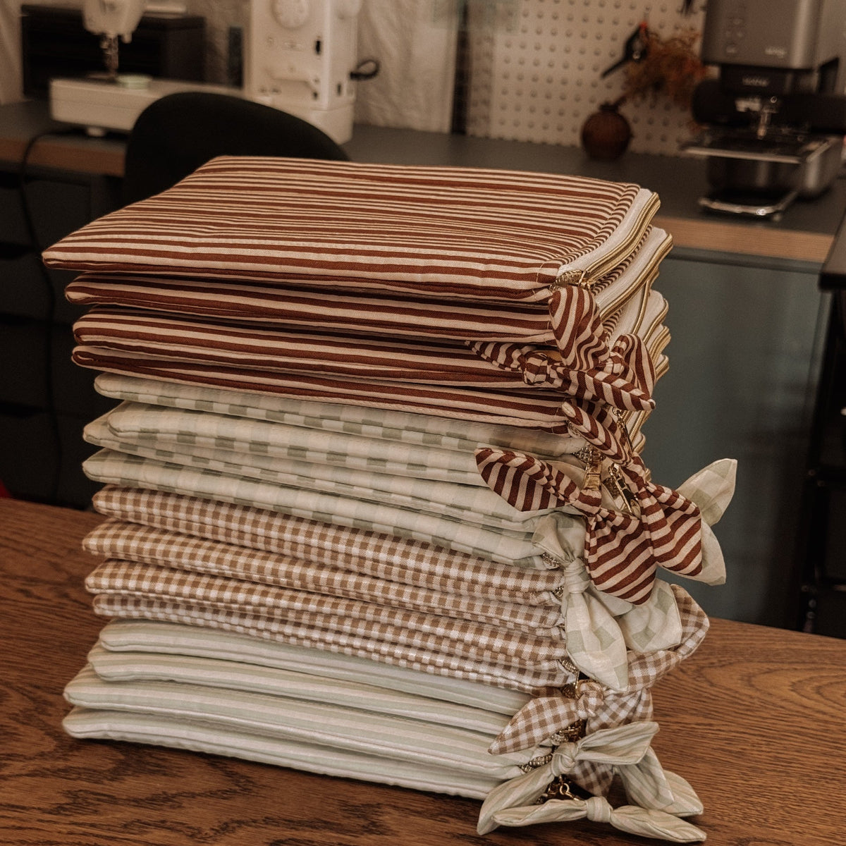 A stack of kindle sleeves items featuring striped and gingham patterns in brown, beige, and white, tied with fabric bows on a wooden table in a sewing room.