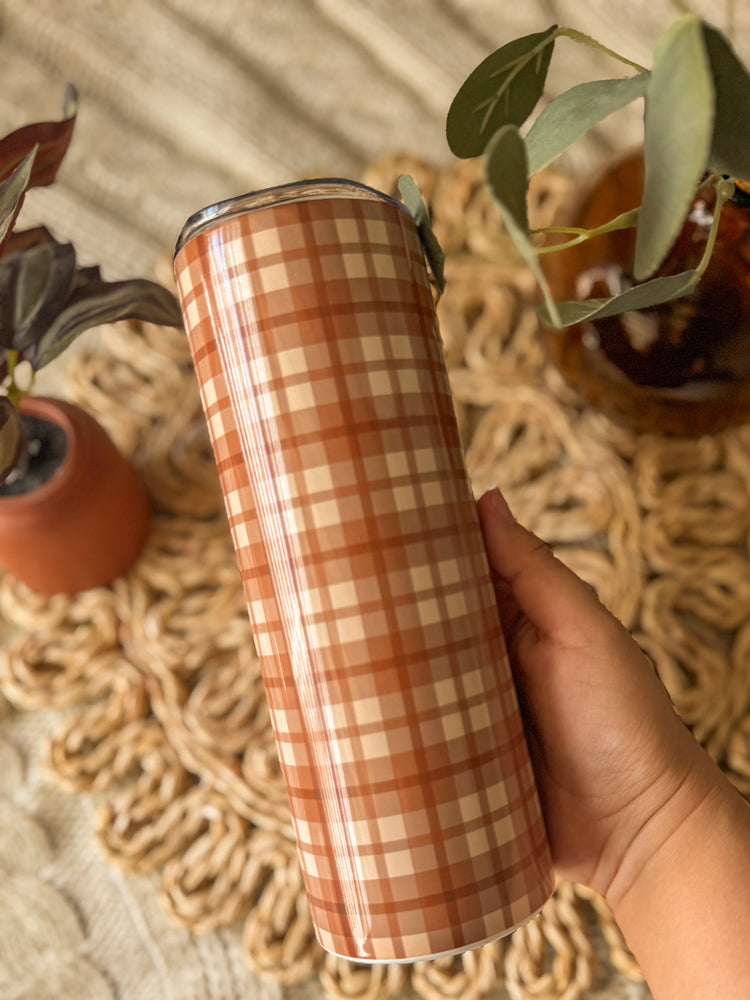 Plaid-patterned tumbler held by a hand on a textured surface with plants.
