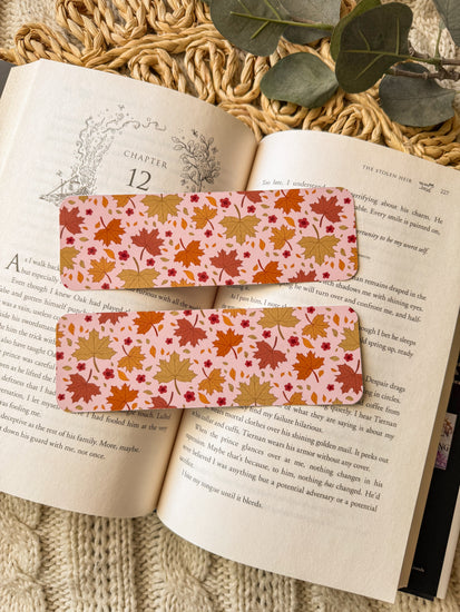 Two autumn-themed bookmarks on an open book with a textured surface and green leaves in the background.