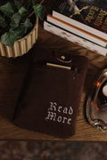 Dark brown book sleeve with 'Read More' text on a wooden surface with books and a plant.