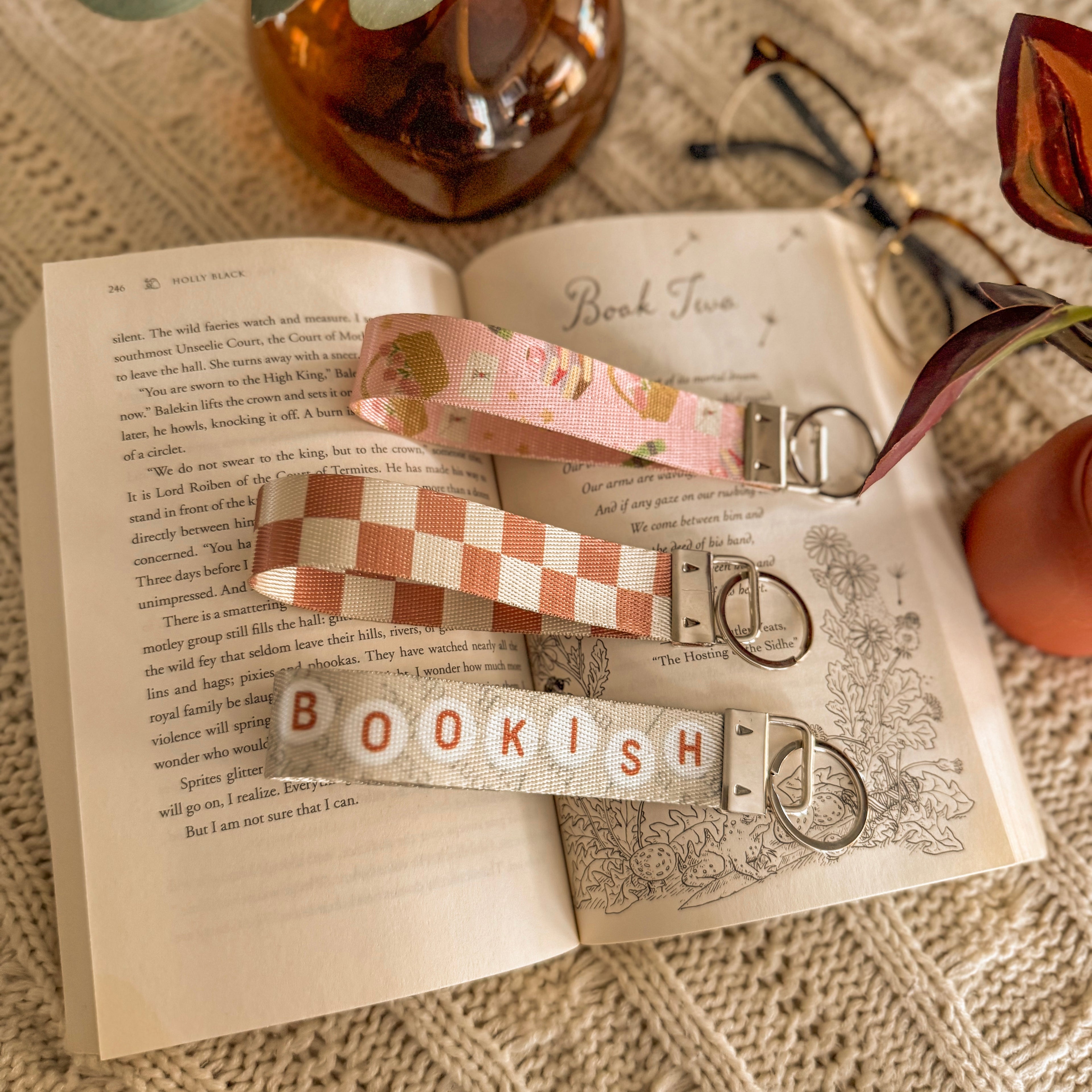 Close up on 3 Bookish Key Fob Wristlets with silver metal detail, on top of an open book