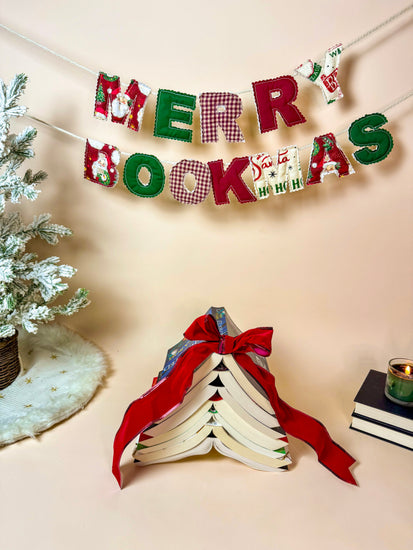 Handmade Merry Bookmas fabric banner hanging above a cozy holiday book scene with a book tree and candle.