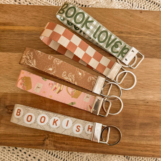Flat lay of 5 styles of bookish key fobs with silver metal hardware on a wood background