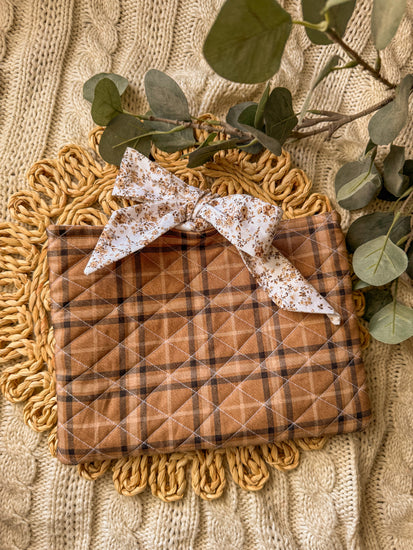 Quilted brown plaid blanket with a white floral bow on a textured surface with green leaves.