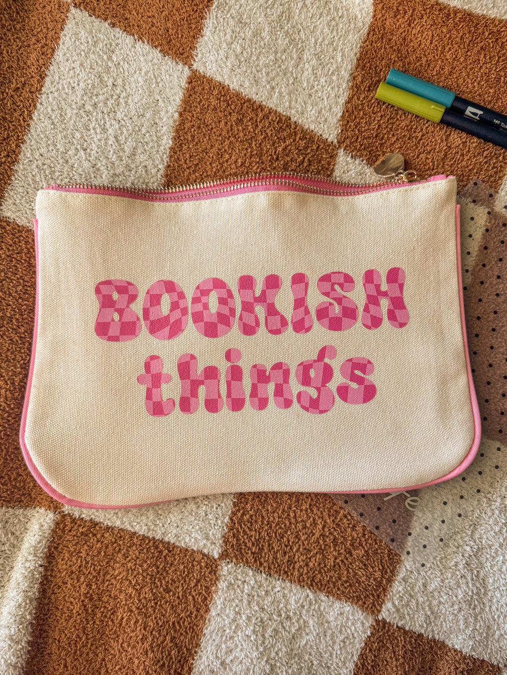 Cream canvas pouch with pink “Bookish Things” text lying on checkered blanket