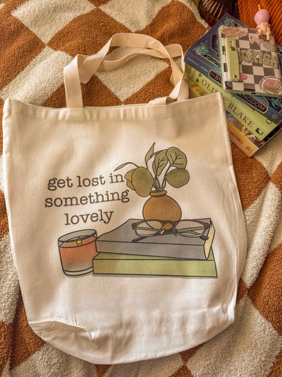 White canvas tote bag with books, glasses, a candle, and plant illustration with the phrase "get lost in something lovely" styled on cozy orange and green bedding.