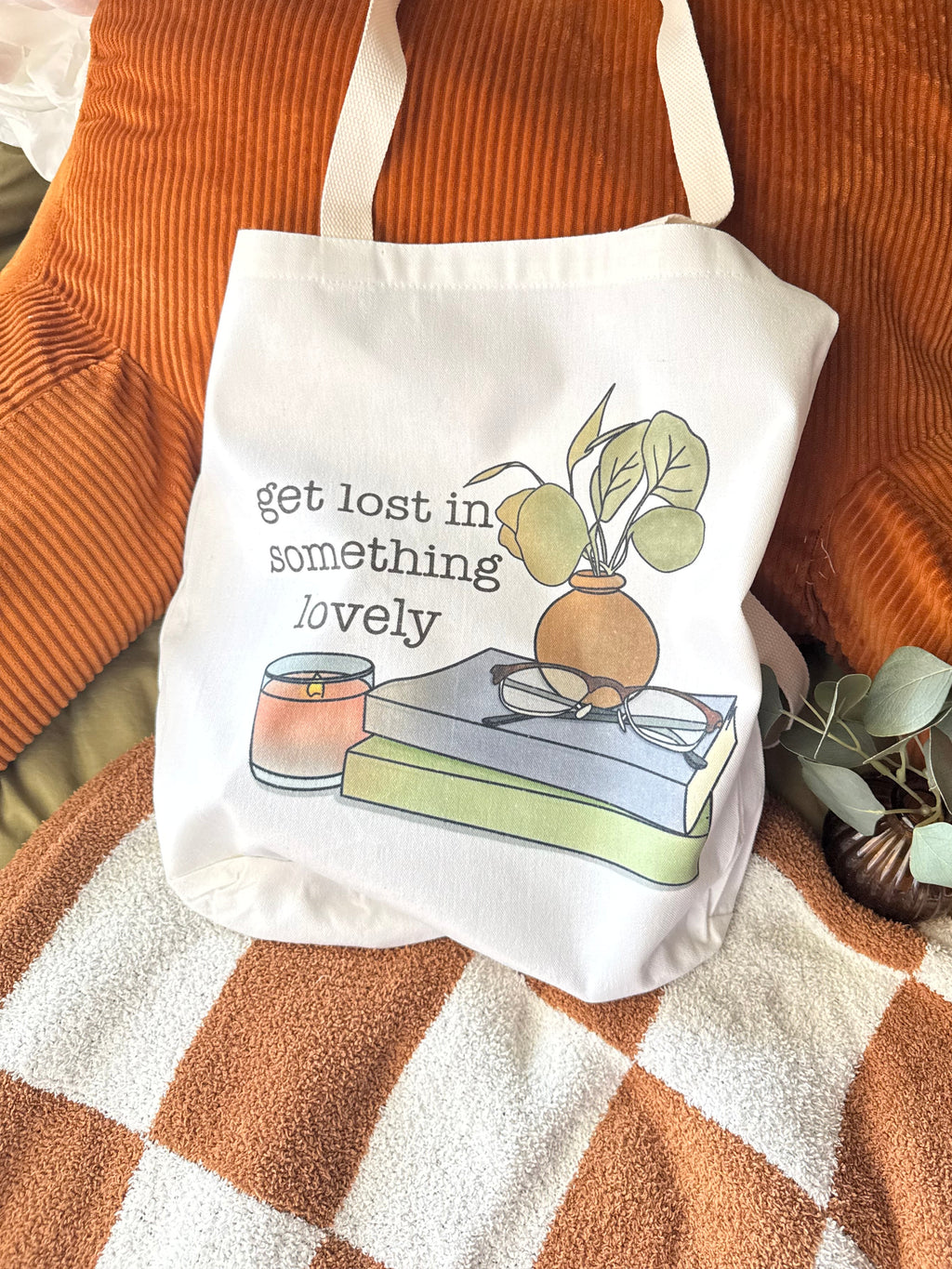 White canvas tote bag with books, glasses, a candle, and plant illustration with the phrase "get lost in something lovely" styled on cozy orange and green bedding.