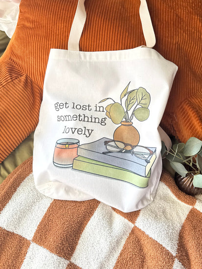 White canvas tote bag with books, glasses, a candle, and plant illustration with the phrase "get lost in something lovely" styled on cozy orange and green bedding.