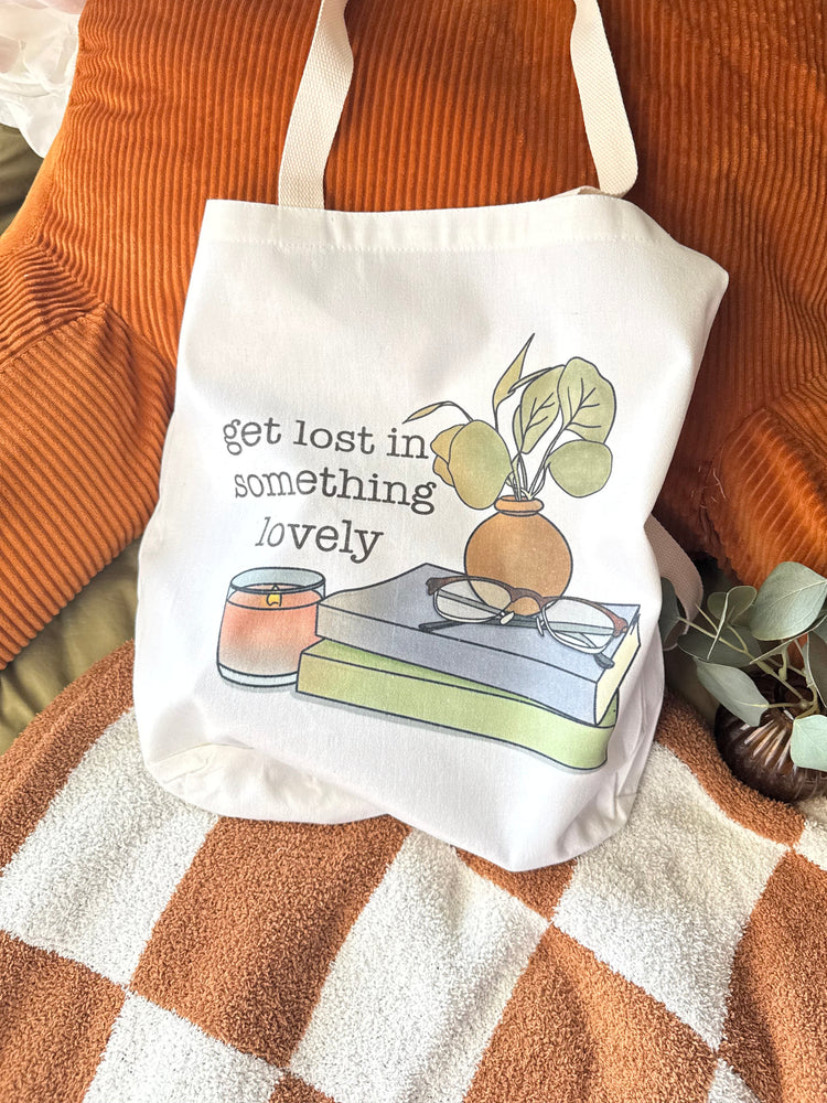 White canvas tote bag with books, glasses, a candle, and plant illustration with the phrase "get lost in something lovely" styled on cozy orange and green bedding.