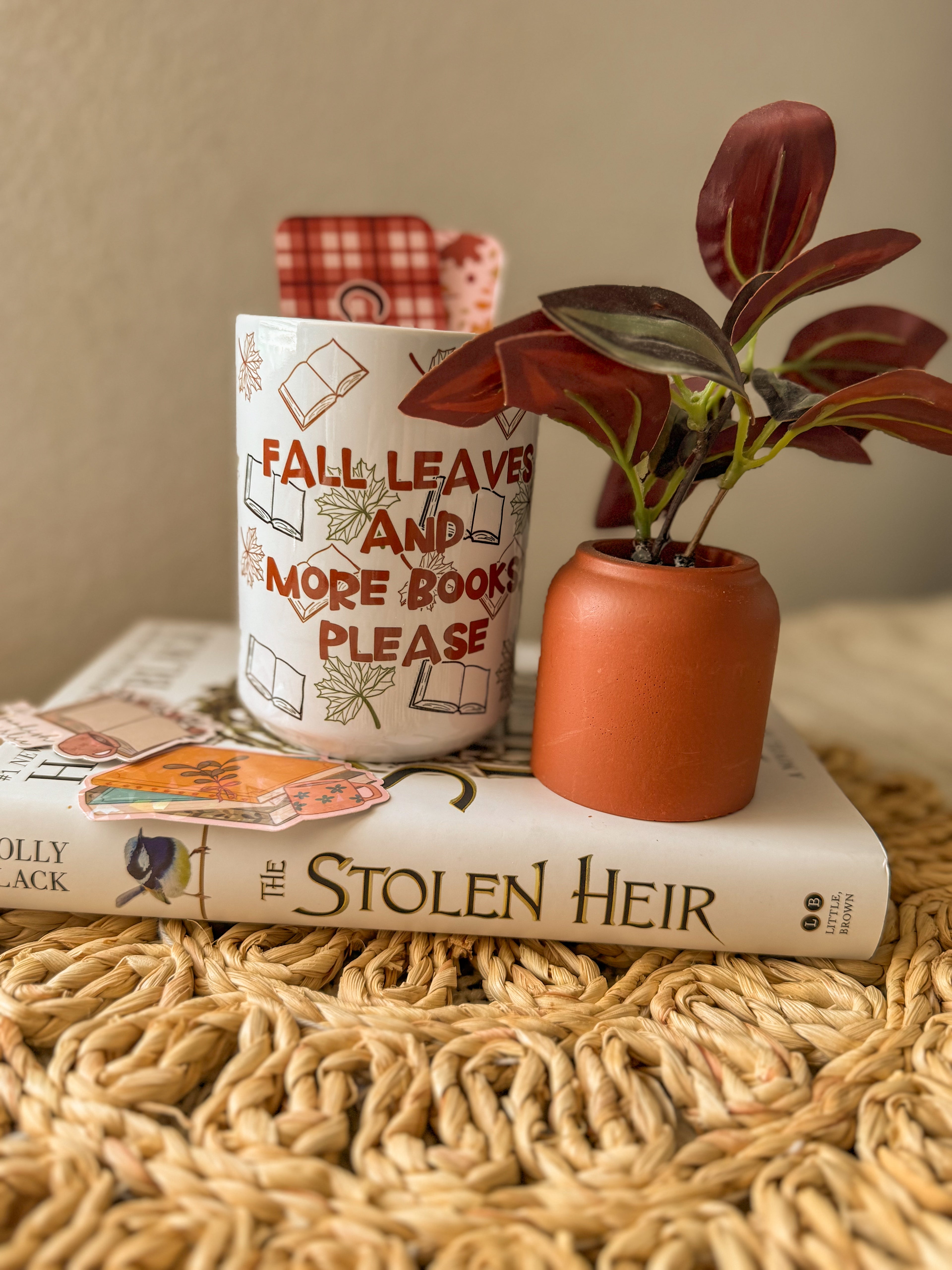 Fall themed coffee cup, book titled 'The Stolen Heir', and small plant on a textured surface.