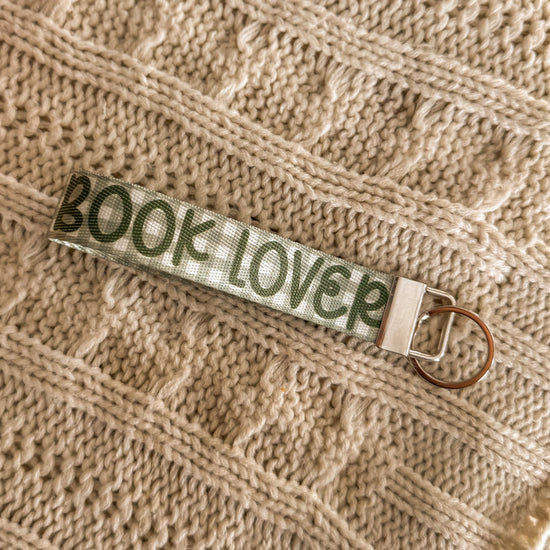 Close up of Green key fob, background is green gingham and the text "BOOK LOVER" in a darker green is written on top. 