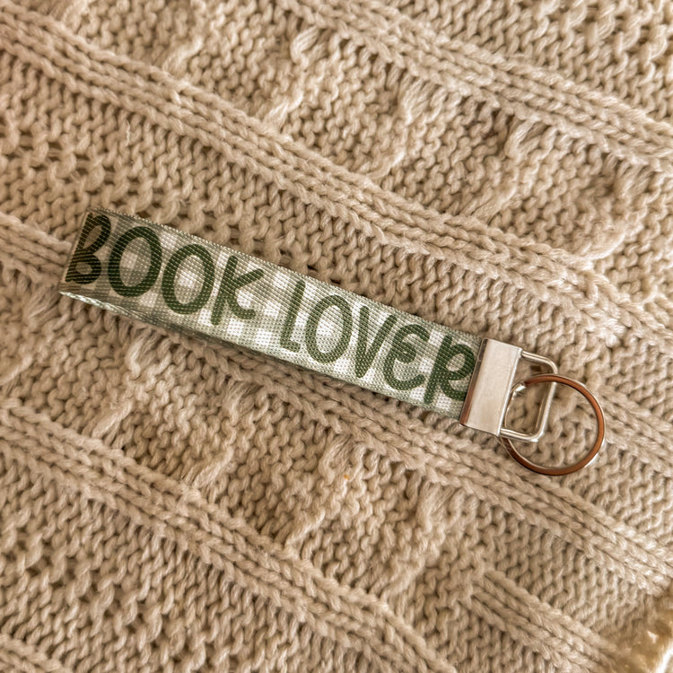 Close up of Green key fob, background is green gingham and the text "BOOK LOVER" in a darker green is written on top. 
