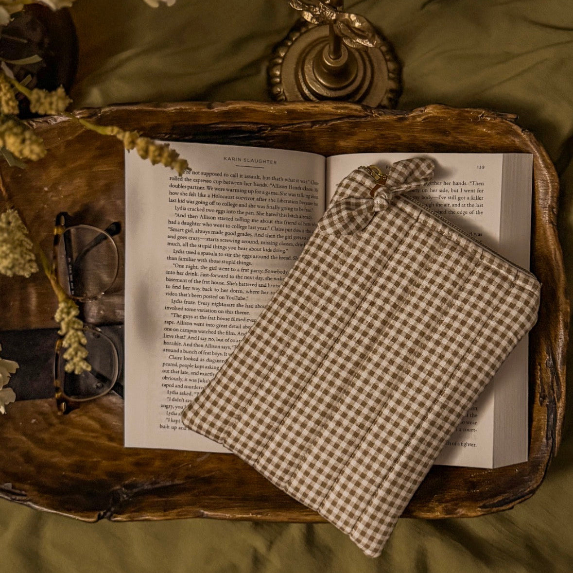 Brown gingham quilted Kindle sleeve with zipper closure resting on an open book, styled on a wooden tray with a candle, glasses, and flowers for a cozy cottagecore reading aesthetic.