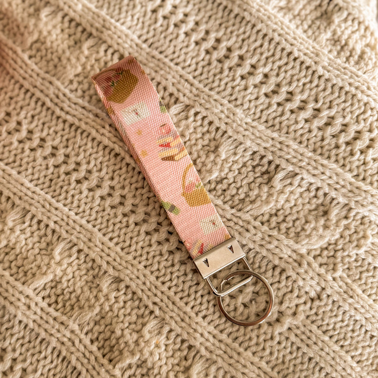 Pink key fob with images of stack of books, picnic baskets, love letters and a wine bottle .