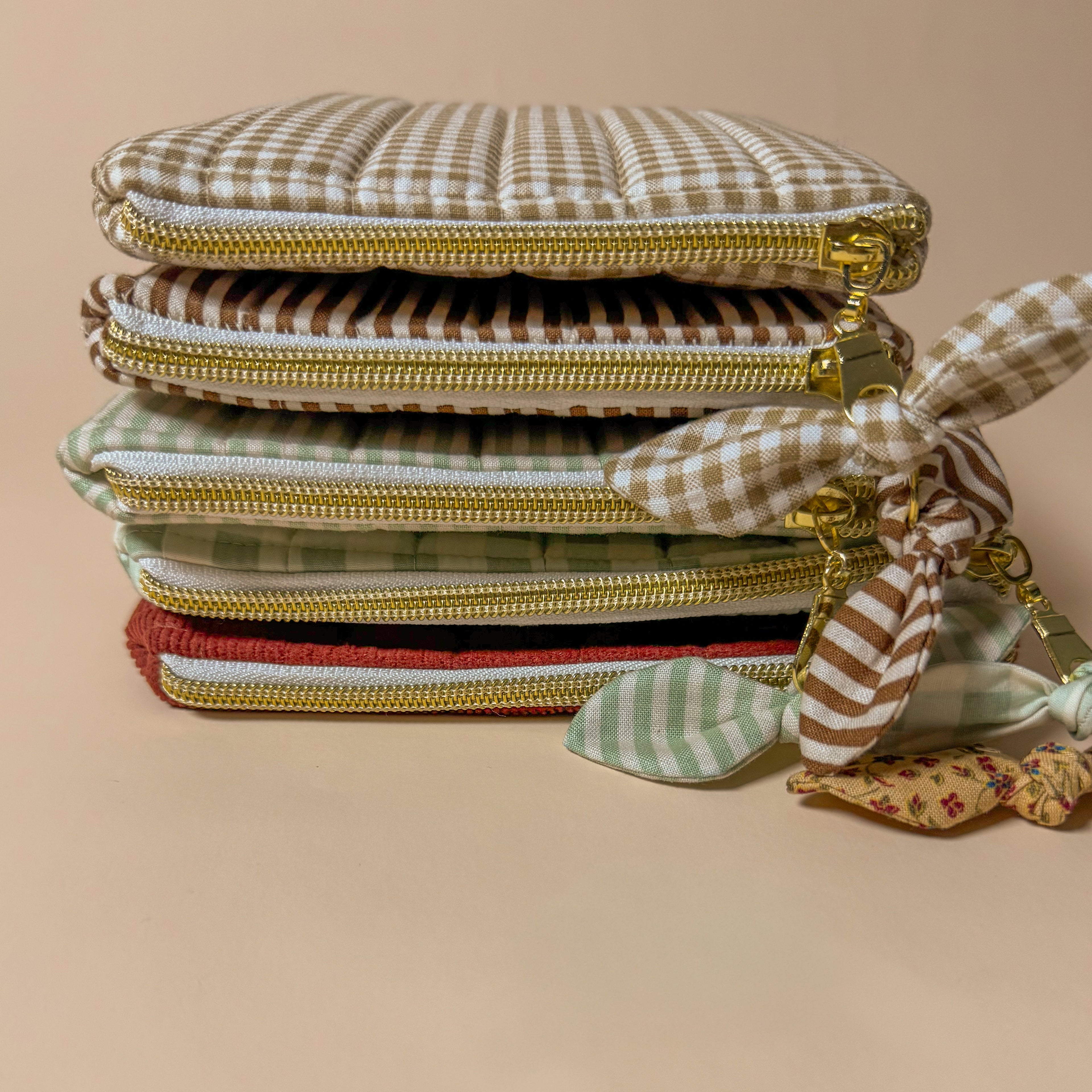 A stack of handmade quilted Kindle sleeves with zippers in various patterns, including brown gingham, sage green checkered, and red corduroy, showing padded e-reader protection for different styles.