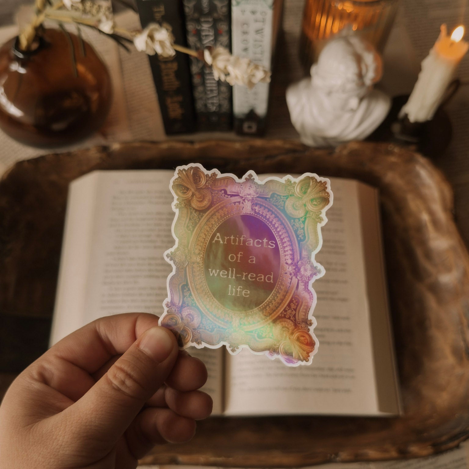 Holographic sticker featuring the quote "Artifacts of a well-read life" in an ornate vintage frame, styled in a cozy Dark Academia setting with books and candles.