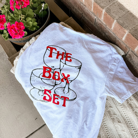 Close-up of graphic tee design showing retro red text “The Box Set” layered over hand-drawn stacked champagne glasses, styled for cozy bookish vibes.