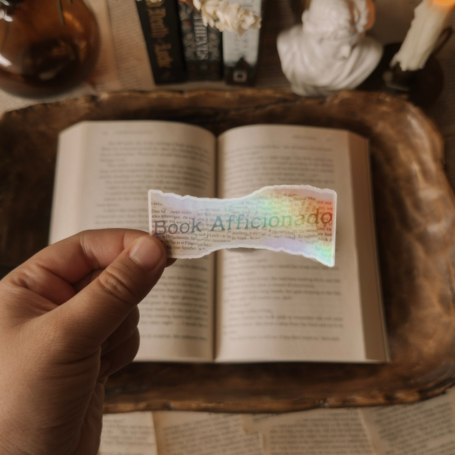 Close-up of a holographic "Book Aficionado" sticker being held over an open book in a dark academia aesthetic setting with candles and vintage books.