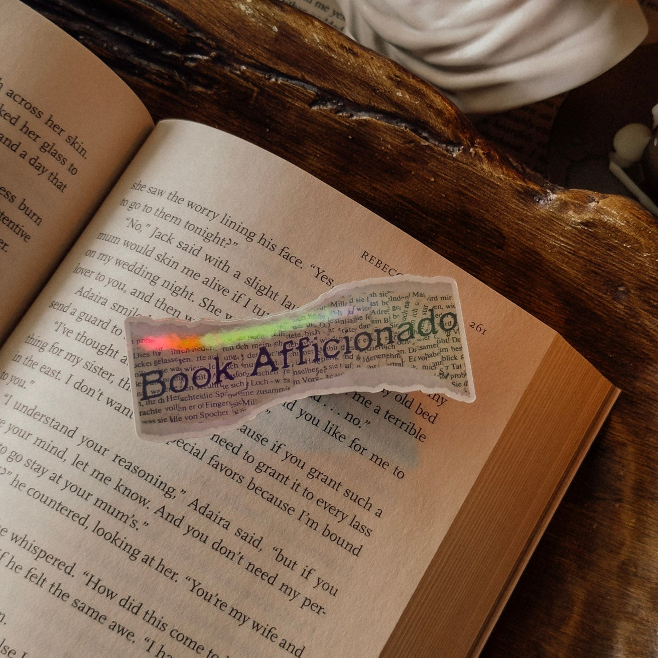 Close-up of a holographic "Book Aficionado" sticker on an open book, showcasing the rainbow shimmer and vintage library aesthetic.