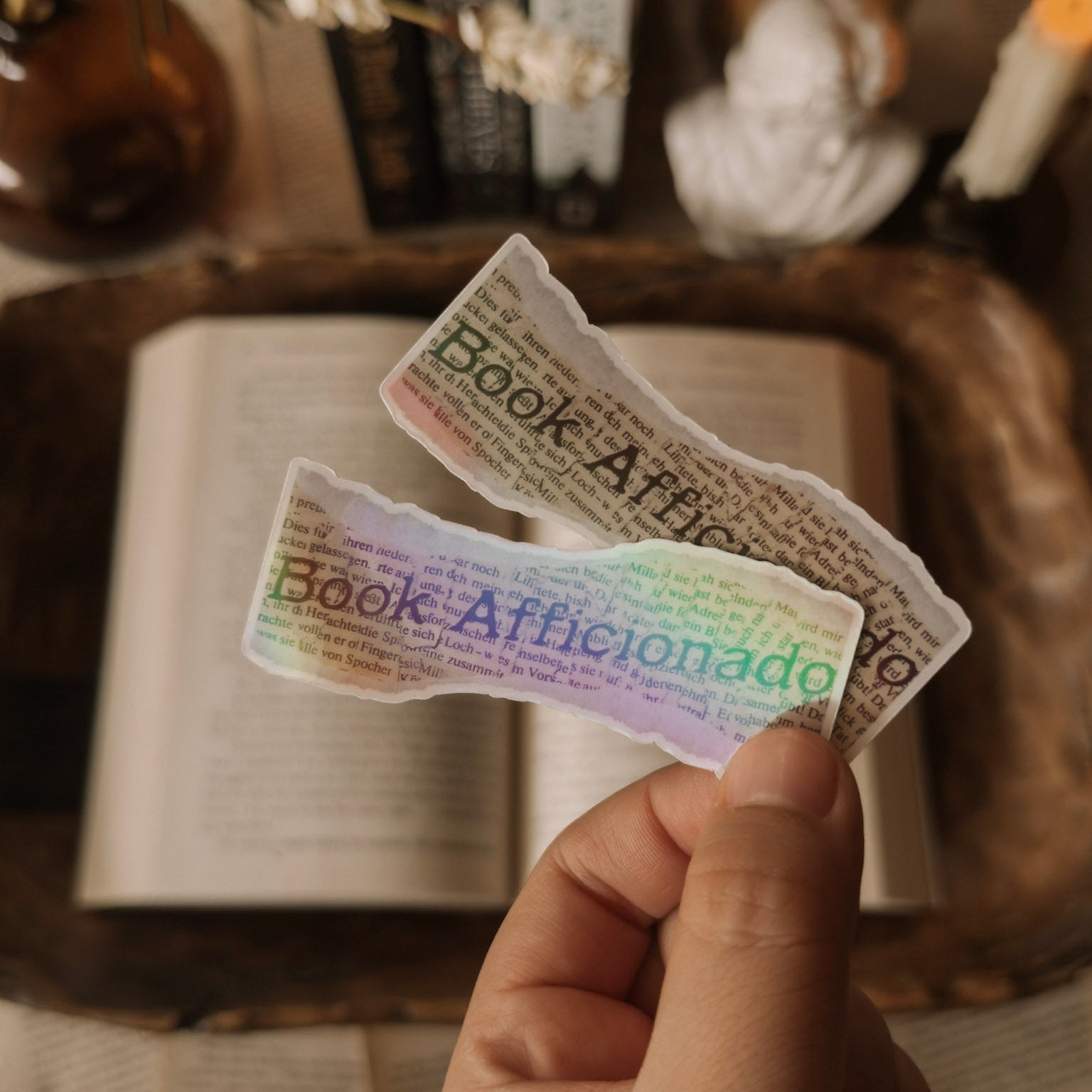 Close-up of a holographic "Book Aficionado" sticker showing the rainbow shimmer, held over an open vintage book with candles in the background.