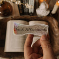 A hand holding a holographic "Book Aficionado" sticker over an open book, showcasing the rainbow shimmer and dark academia aesthetic background.