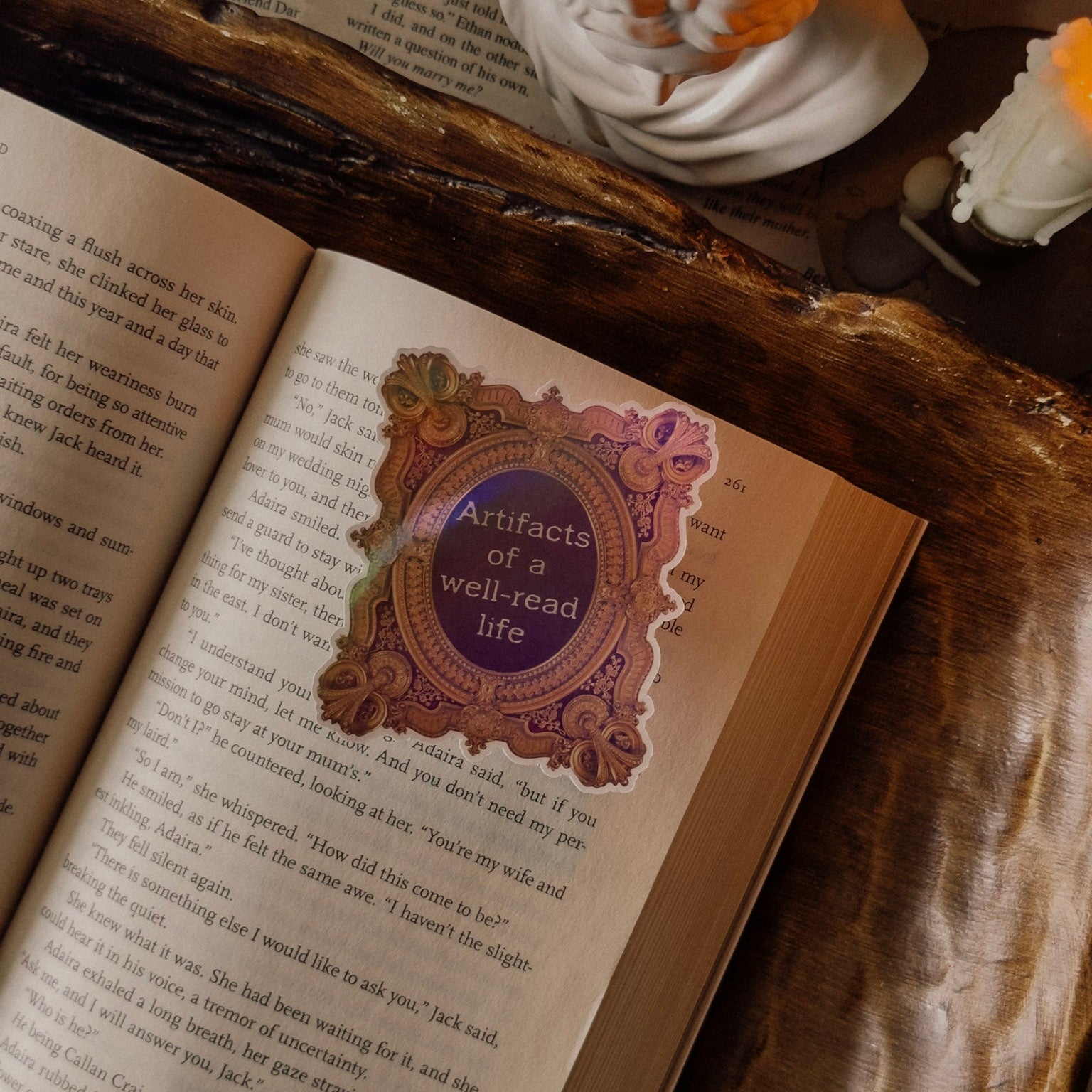 Ornate holographic "Artifacts of a well-read life" sticker placed on an open vintage book, surrounded by dark academia desk decor and a warm candle light.