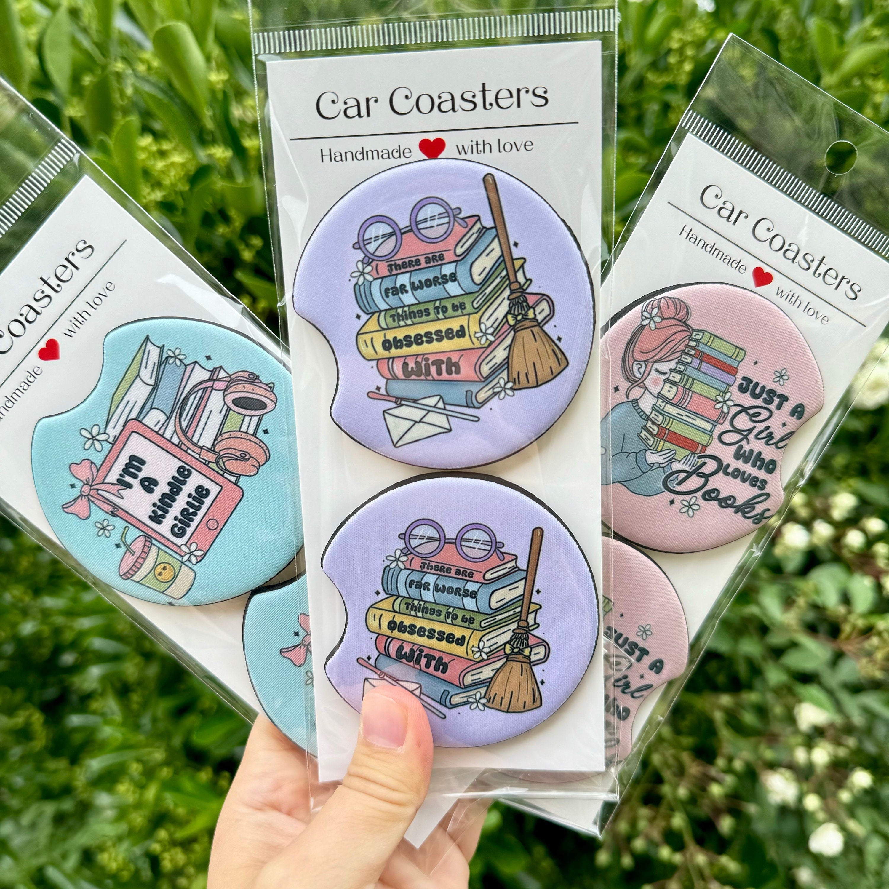 A collection of three Bookish Girlies Car Coasters sets featuring 'Reader Girl', 'Kindle Girlie', and book stack designs, displayed against a natural green background.
