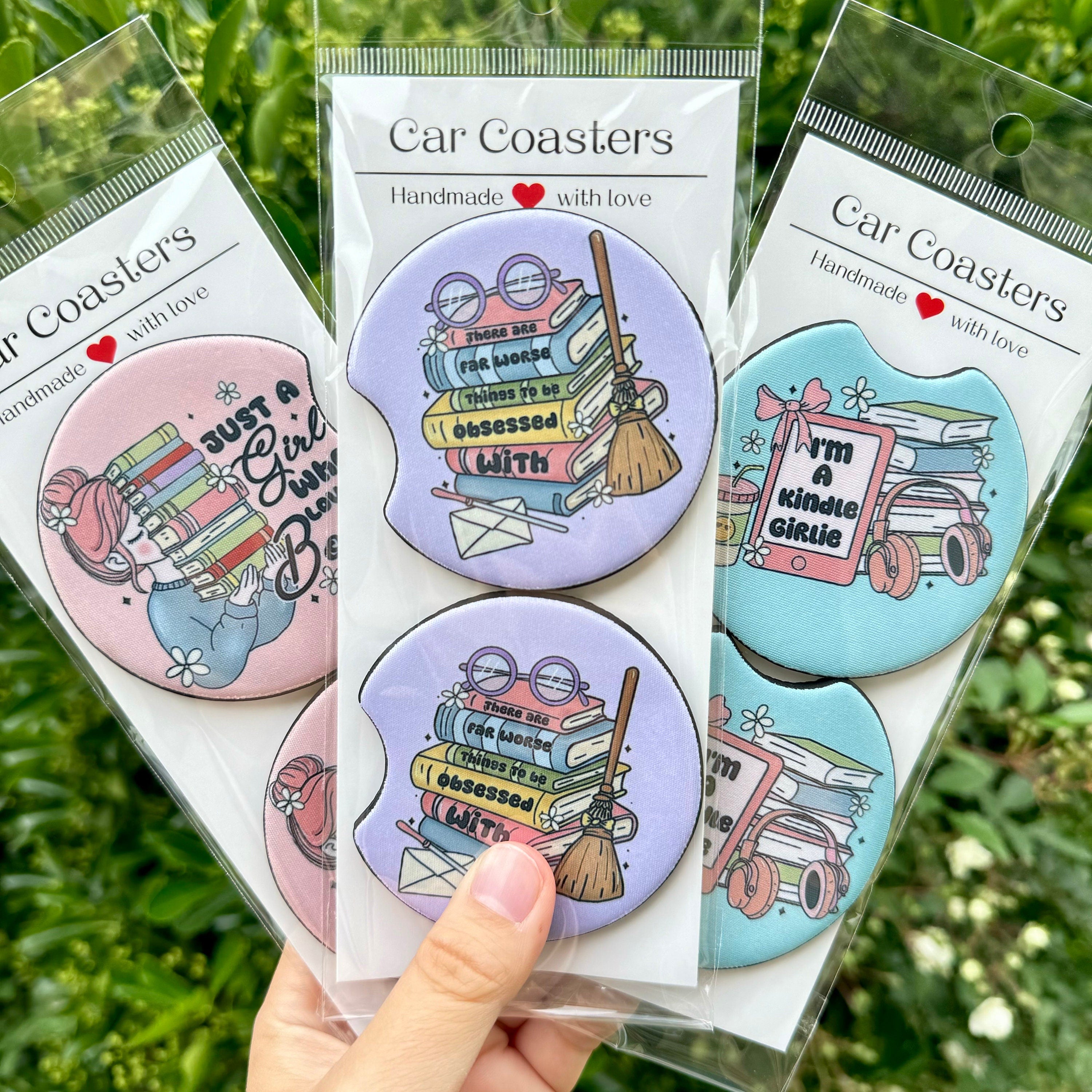 A flat lay of three Bookish Girlies Car Coaster packs by Dex Avenue, featuring 'Reader Girl' (pink), 'Stack of Books' (purple), and 'Kindle Girlie' (blue) designs against a green leafy background