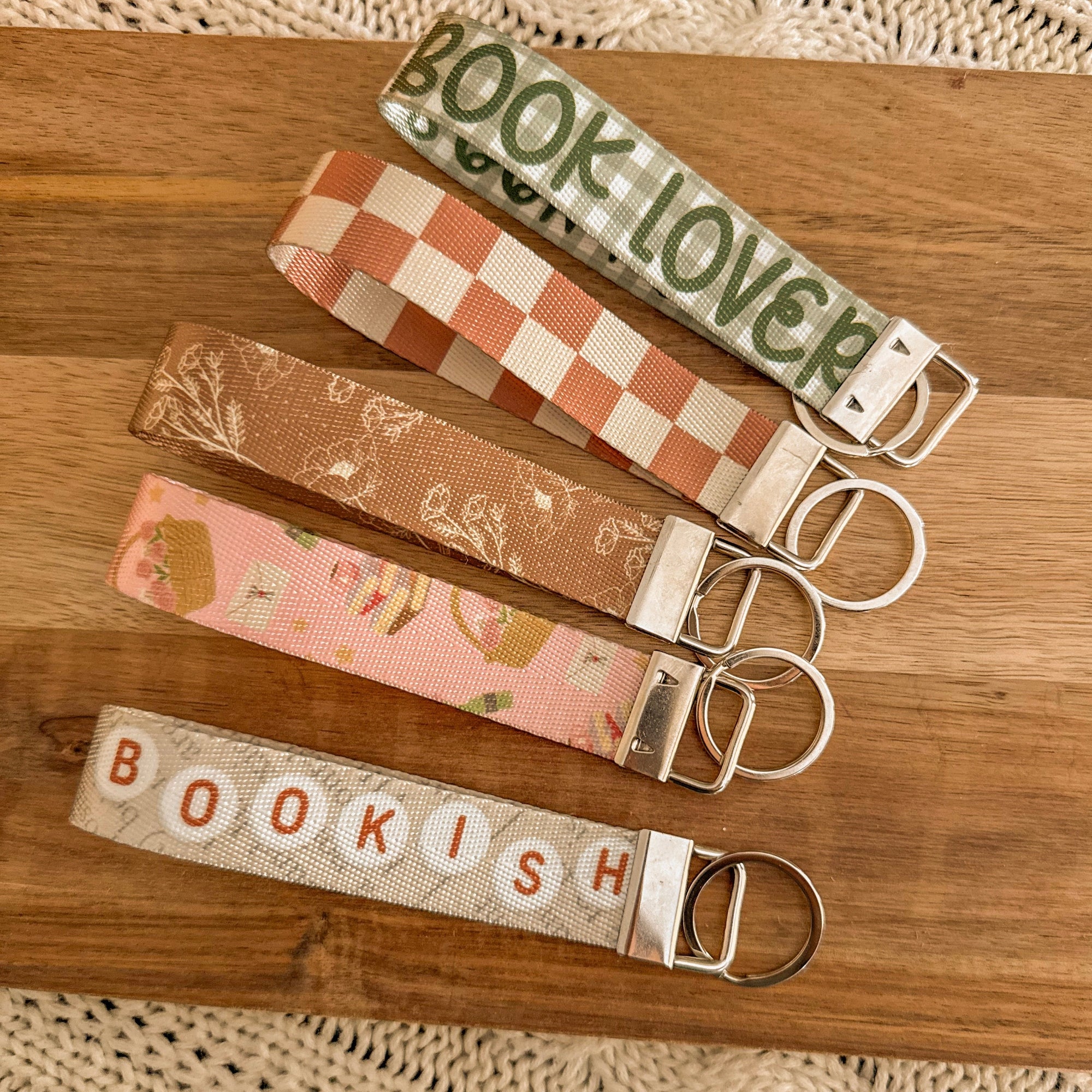 A flat lay of five handmade bookish key fobs in checkered, floral, and 'Book Lover' patterns. The wristlet keychains feature silver hardware and are styled on a warm wood background, perfect for a cozy reader's aesthetic.