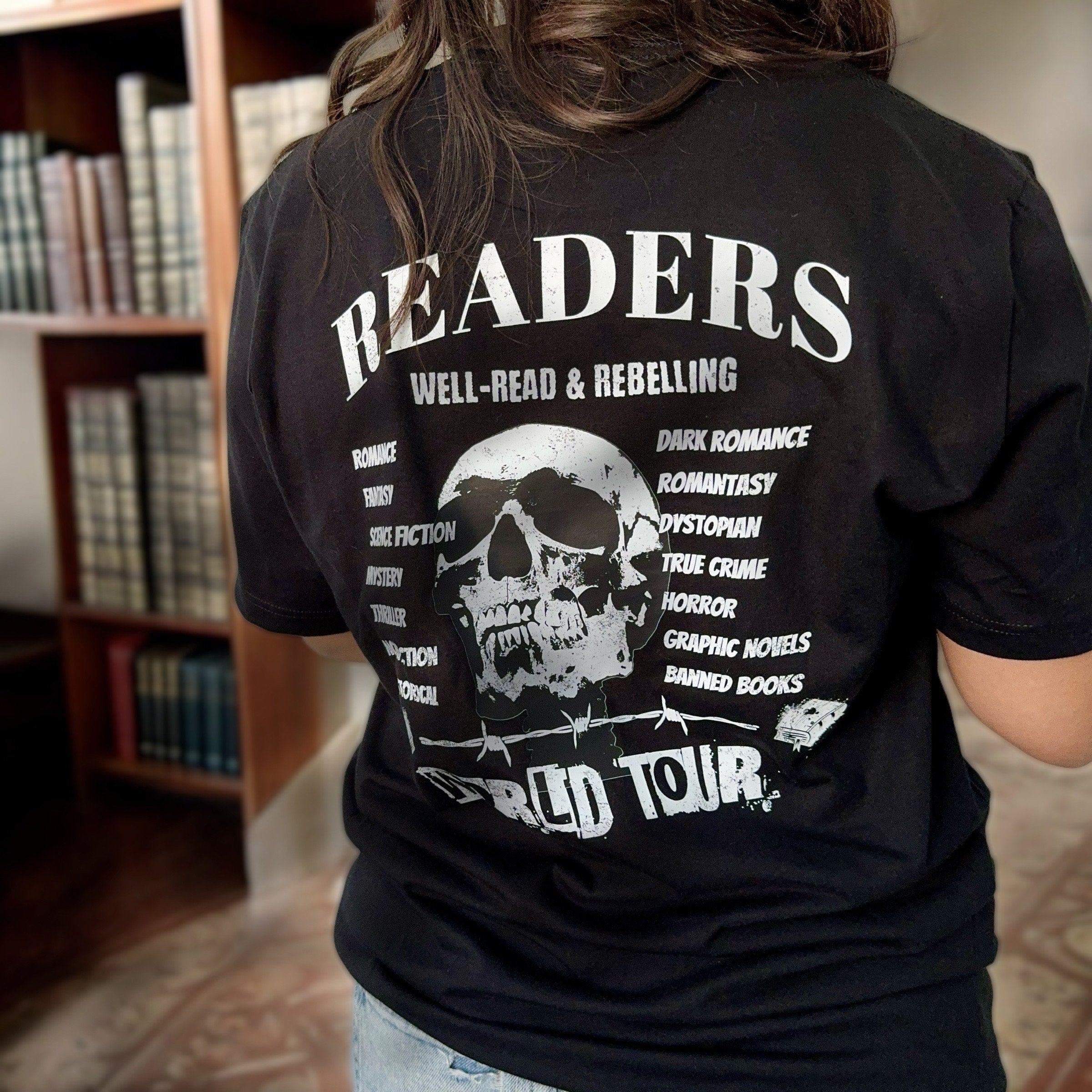 Back view of the Bookish World Tour Tee by Dex Avenue, featuring a white skull graphic and book genres list on a black t-shirt, styled in a library setting.