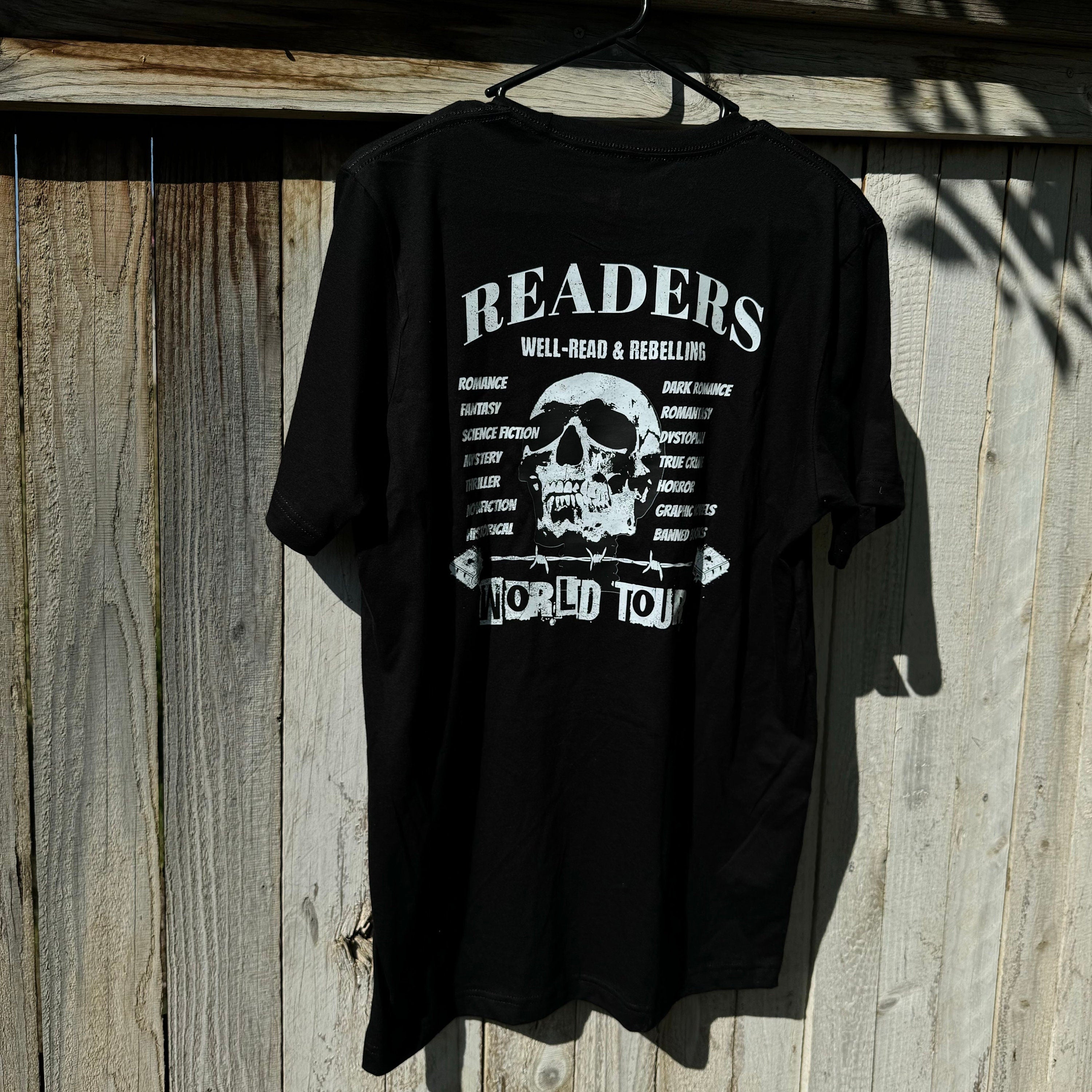 Back view of the black Bookish World Tour Tee by Dex Avenue, featuring a white skull graphic and book genres list, hanging on a wooden fence in natural sunlight.