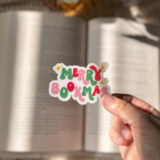 Hand holding a Merry Bookmas vinyl sticker over an open book with warm lighting.