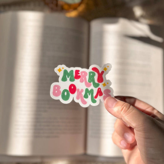 Hand holding a Merry Bookmas vinyl sticker over an open book with warm lighting.