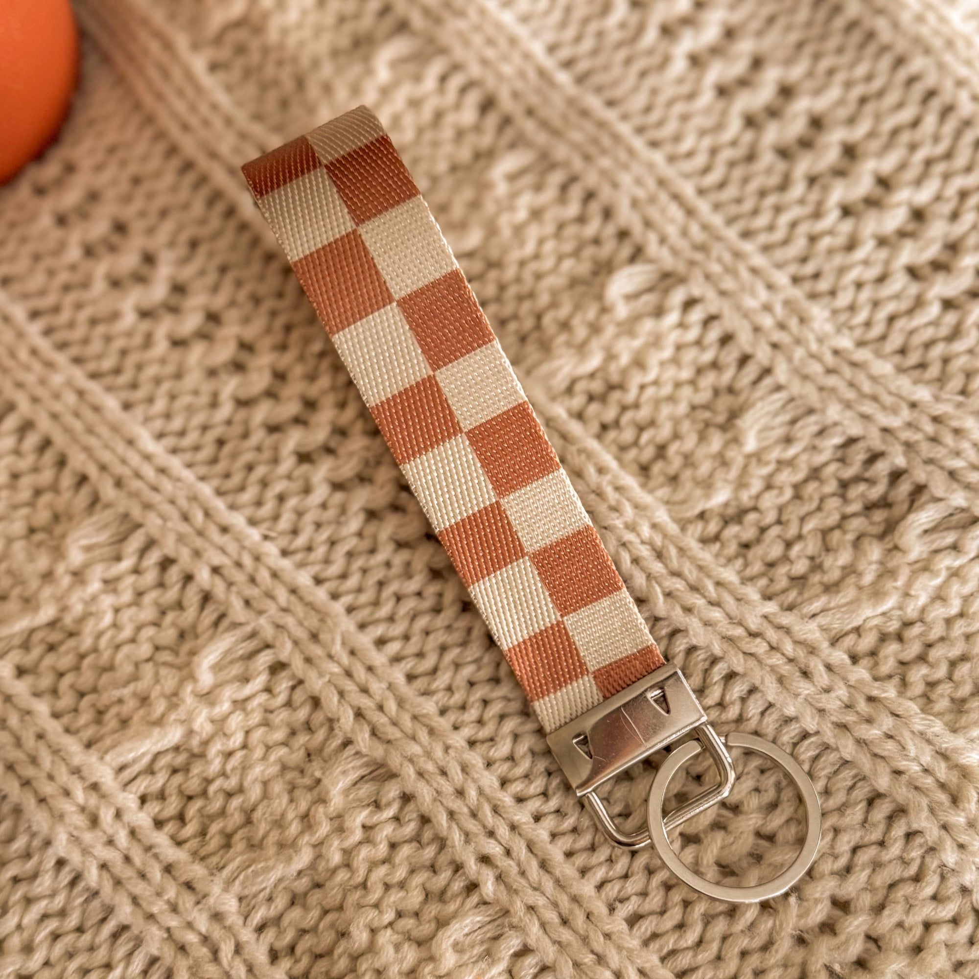 A neutral brown and cream checkered fabric key fob with a silver metal clasp, styled on a warm tan cable knit blanket. This handcrafted wristlet accessory is designed for a cozy, bookish lifestyle.